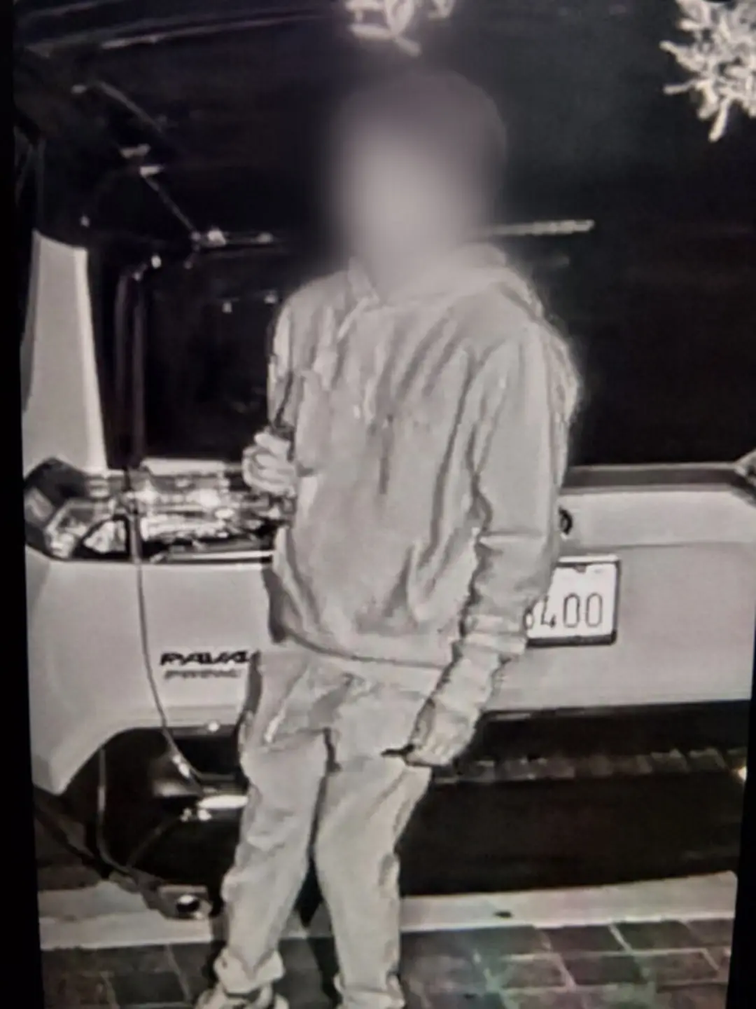 A person in a hoodie and pants stands in front of a parked SUV in a dimly lit area, holding an object in their right hand.